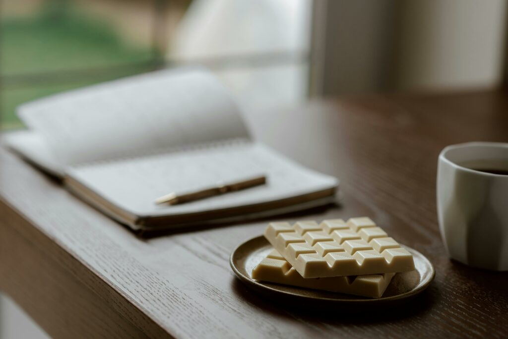 White chocolate on ceramic plate near opened notebook with pen and cup of coffee